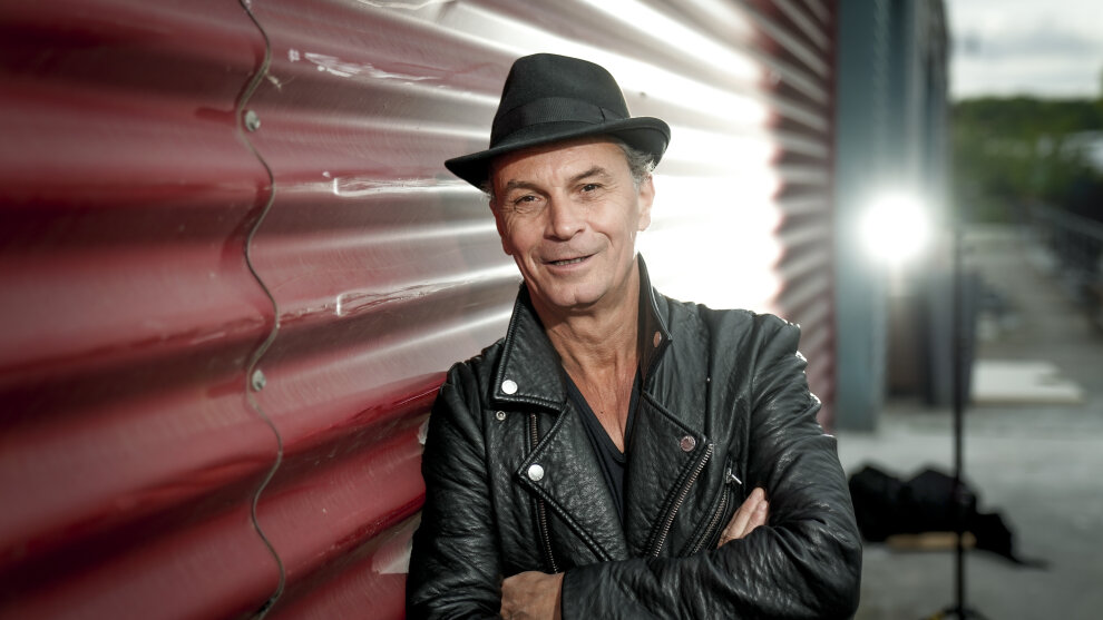 Das Foto zeigt den Ehrenamtspaten 2026 Peter Brings in schwarzer Lederjacke und mit schwarzem Hut, der sich an eine Mauer anlehnt. Im Hintergrund ist ein Scheinwerfer zu sehen, der Peter Brings von hinten anleuchtet.