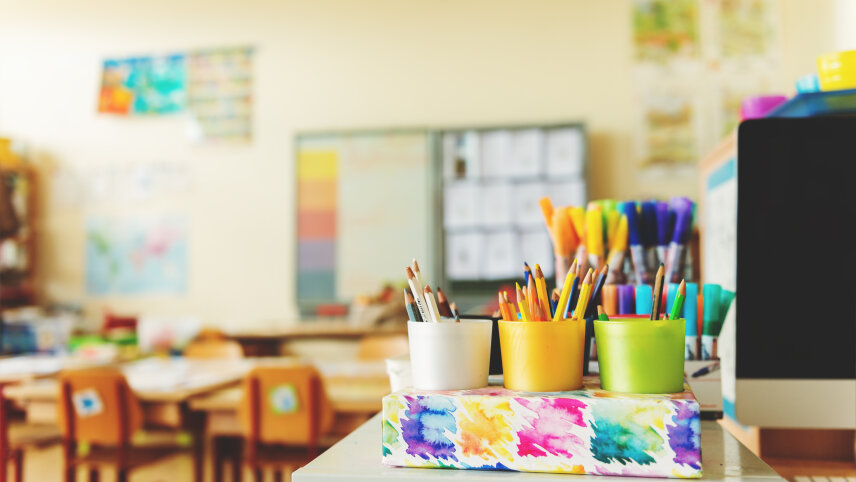 Blick in einen Klassenraum. Im Vordergrund stehen mehrere Becher mit Farbstiften. Im Hintergrund sind kleine Stühle und Kreidetafeln zu sehen.