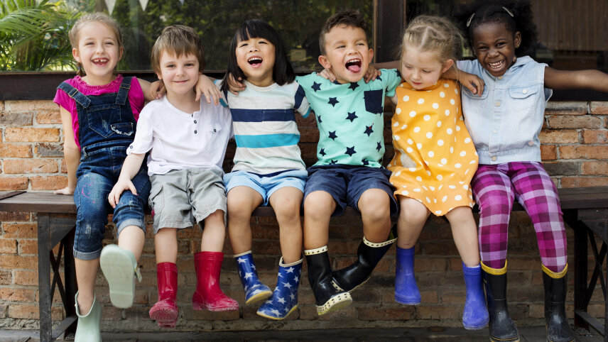 Eine Gruppe von Kindergartenkindern sitzt Arm in Arm auf einer Bank. Alle Kinder schauen fröhlich und lachen., © Smarterpix/Rawpixel