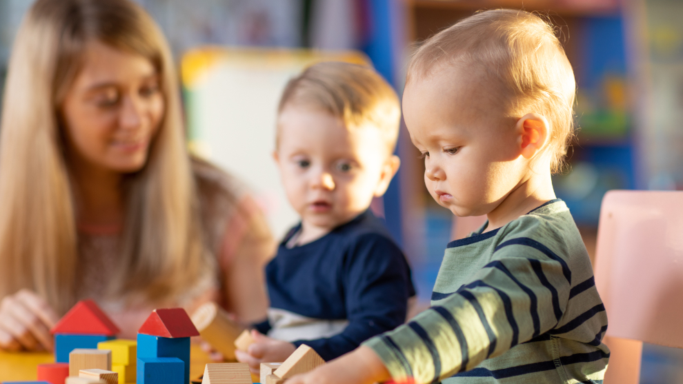 Junge blonde Frau spielt mit zweit kleinen Kindern. Auf dem Tisch liegen bunte Bauklötze aus Holz.
