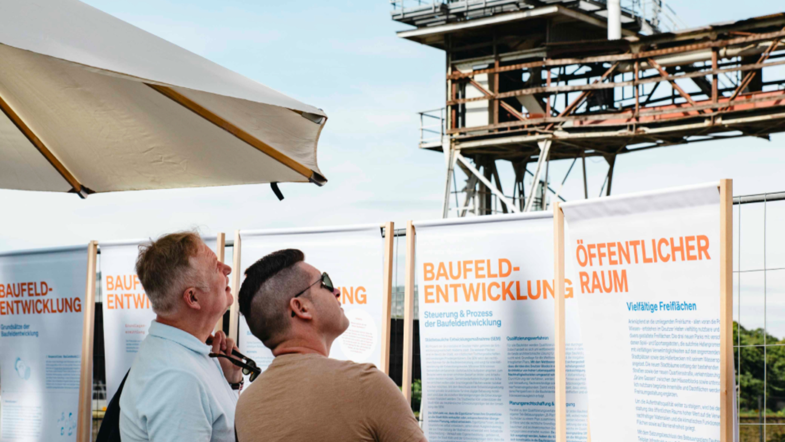Zwei Besucher des Hafentages 2023 stehen im Freien unter Sonnenschirmen im Biergarten der Essigfabrik und betrachten auf die Abbruchkante der Ellmühle. In der Mitte des Bildes stehen Ausstellungs-Roll-Ups mit Informationen zum Öffentlichen Raum und Baufeldentwicklung im Deutzer Hafen. Im Hintergrund ist ein Industriekran zu sehen.