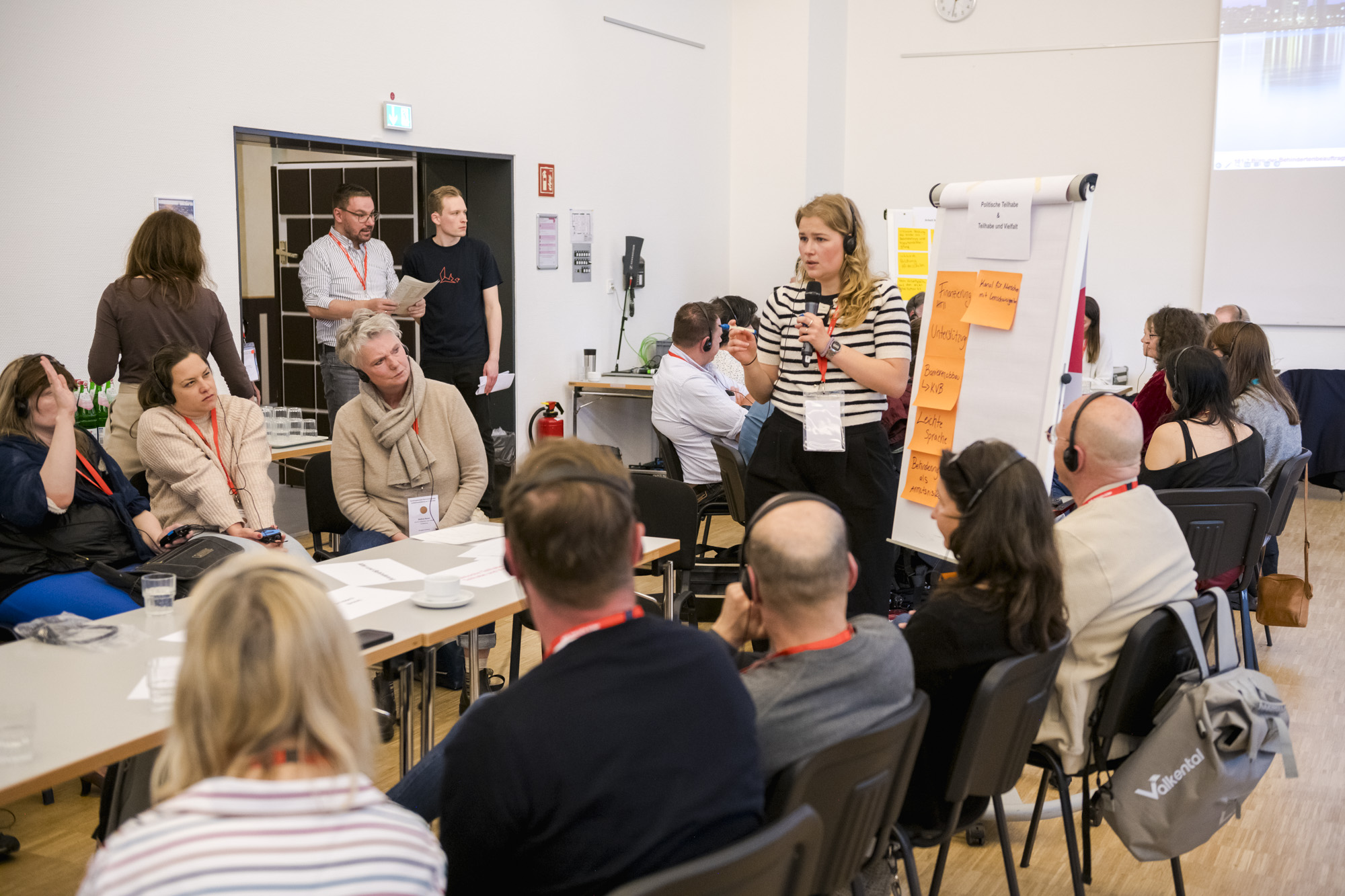 Eine Person steht am Flip-Chart an einem Tisch, weitere Personen sitzen an einem Tisch. Drei Personen stehen an der Tür.