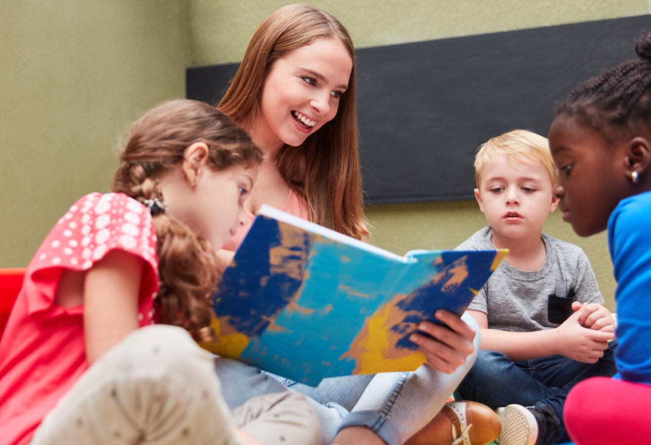 Erzieherin liest aus einem spannenden Kinderbuch vor im Kindergarten oder einer Kita, © © adobe.stock.com - Robert Kneschke