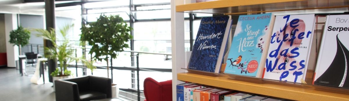 Im Vordergrund ein Buchregal mit mehreren Büchern ausgestellt, dahinter zwei Sessel, © Stadtbibliothek Köln