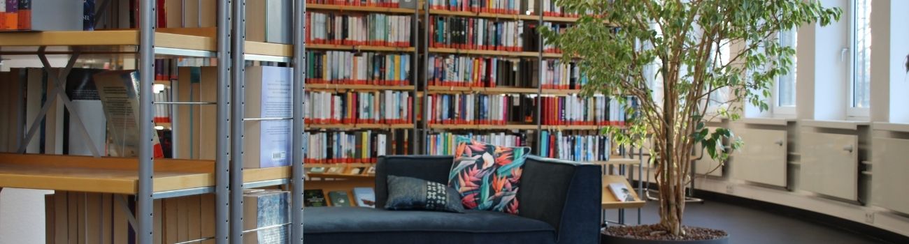 vorderer Bereich ein Teil des Bücherregals, dahinter Liegesofa vor weiterem Bücherregal mit einer Pflanze daneben, © Stadtbibliothek Köln