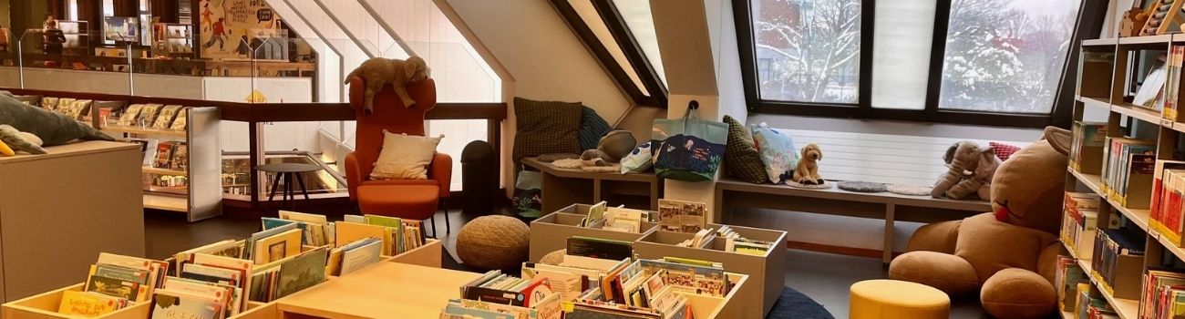 Kinderbereich der Stadtteilbibliothek Porz, ein orangener Ohrensessel, viele Stofftiere, Tröge mit Kinderbüchern im Vordergrund, rechts ein Regal, daneben ein großer Stoffhase, © Stadtbibliothek Köln