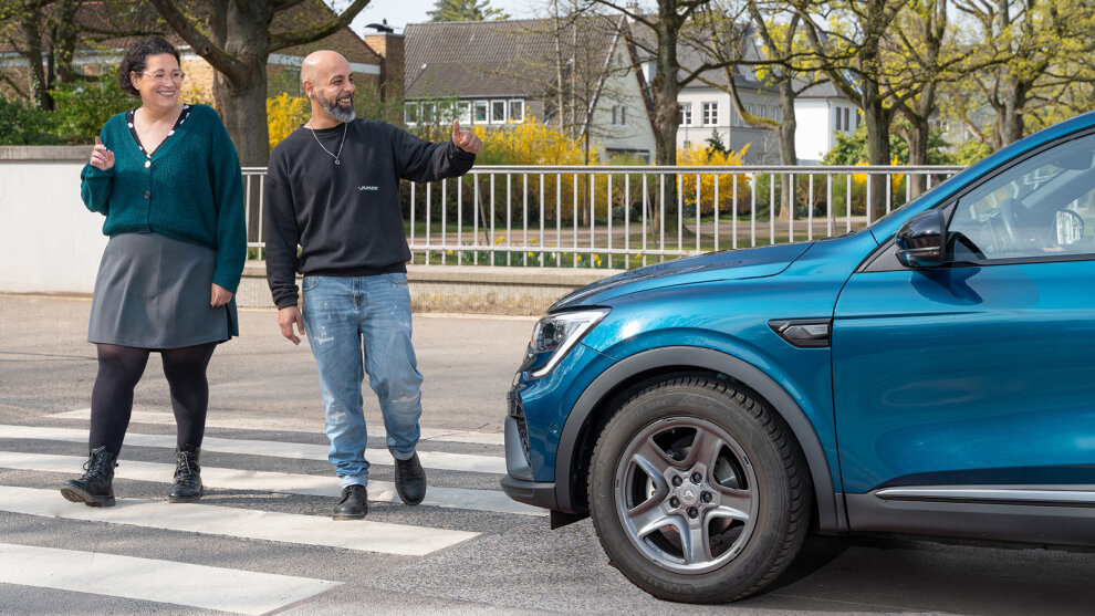 Zwei zu Fuß Gehende queren die Straße über einen Zebrastreifen. Ein Auto hat für sie angehalten. Sie bedanken sich mit einem Lächeln.
