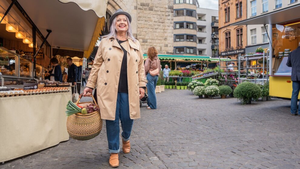 Plakatmotiv der Aktion "Stadt, Land, zu Fuß". Eine Frau mit Hut, Mantel und Einkaufskorb voller Gemüse geht über einen Kölner Wochenmarkt.