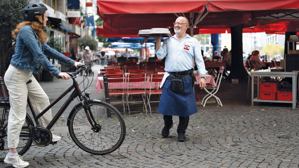 Am linken Bildrand ist eine Radfahrerin zu sehen, die ein Bein am Boden hat. Rechts neben ihr steht ein Köbes mit einem Tablett in der Hand, der ihr freundlich zulächelt.