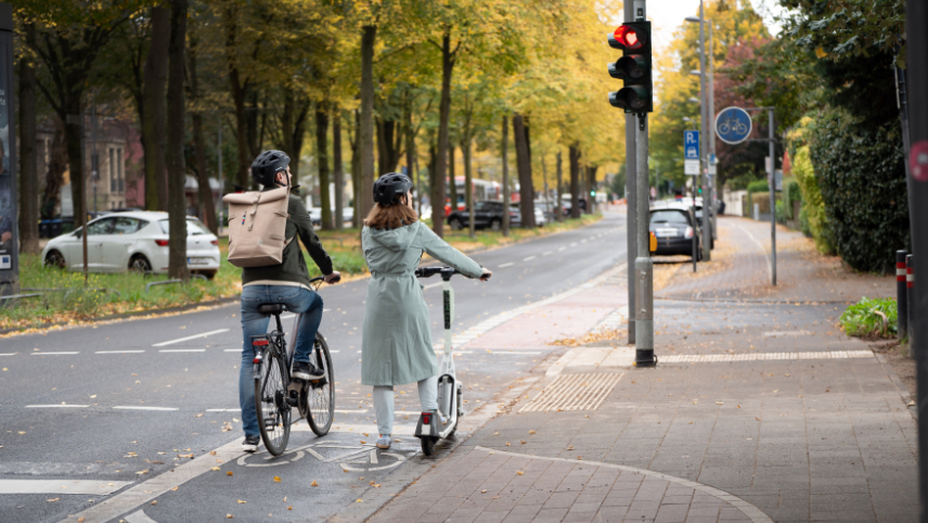 Zu sehen sind ein Mann auf einem Fahrrad und eine Frau auf einem E-Scooter, die nebeneinander an einer roten Ampel warten. Das Rotsignal der Ampel ist in Herzform.